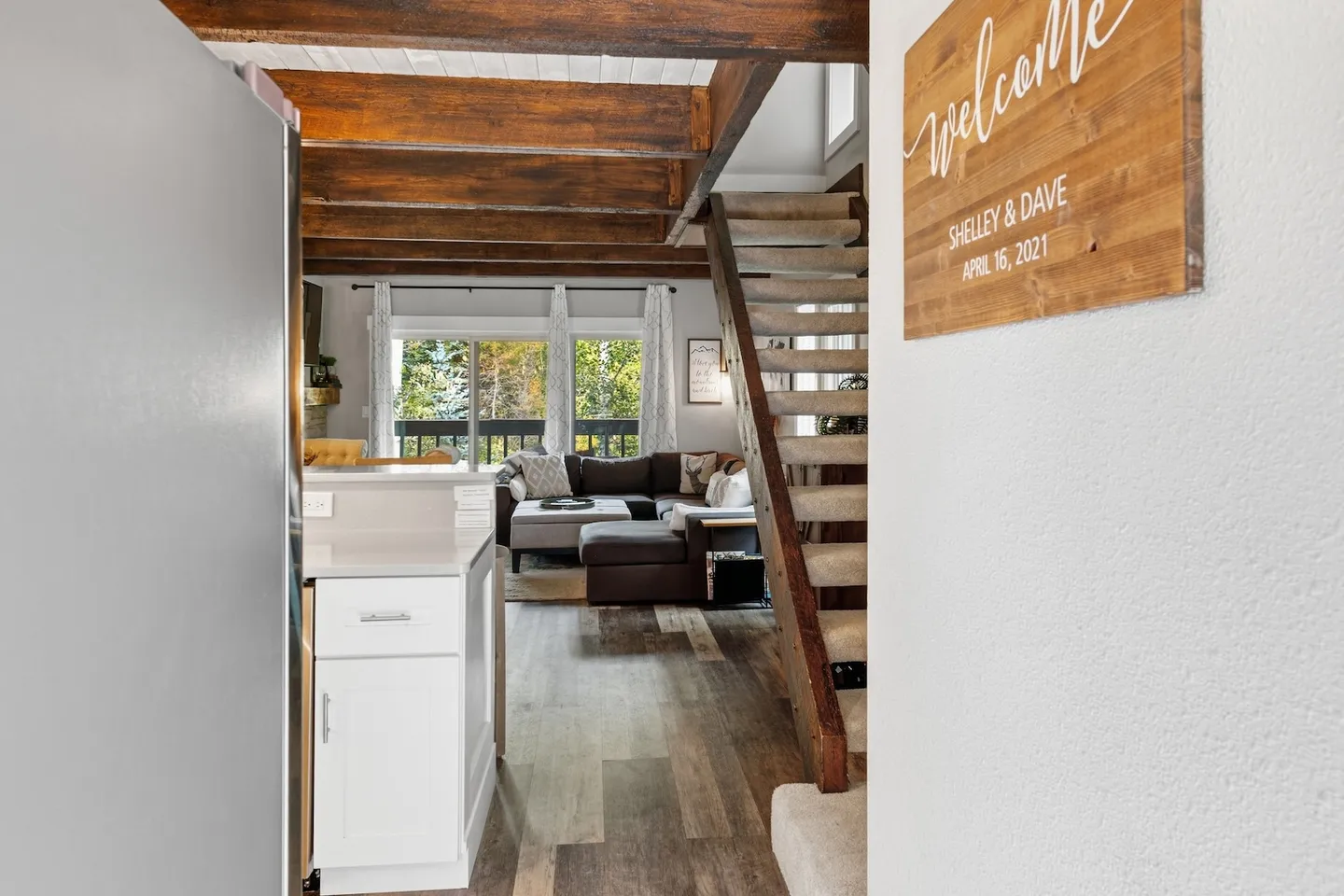 View from kitchen entry past carpeted stairs leading toward an open living room with exposed ceiling beams and sliding glass doors.