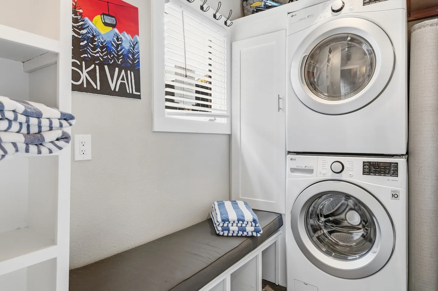 A laundry nook and mudroom area with a stacked washer and dryer, a cushioned bench, folded striped towels, and a Ski Vail poster.
