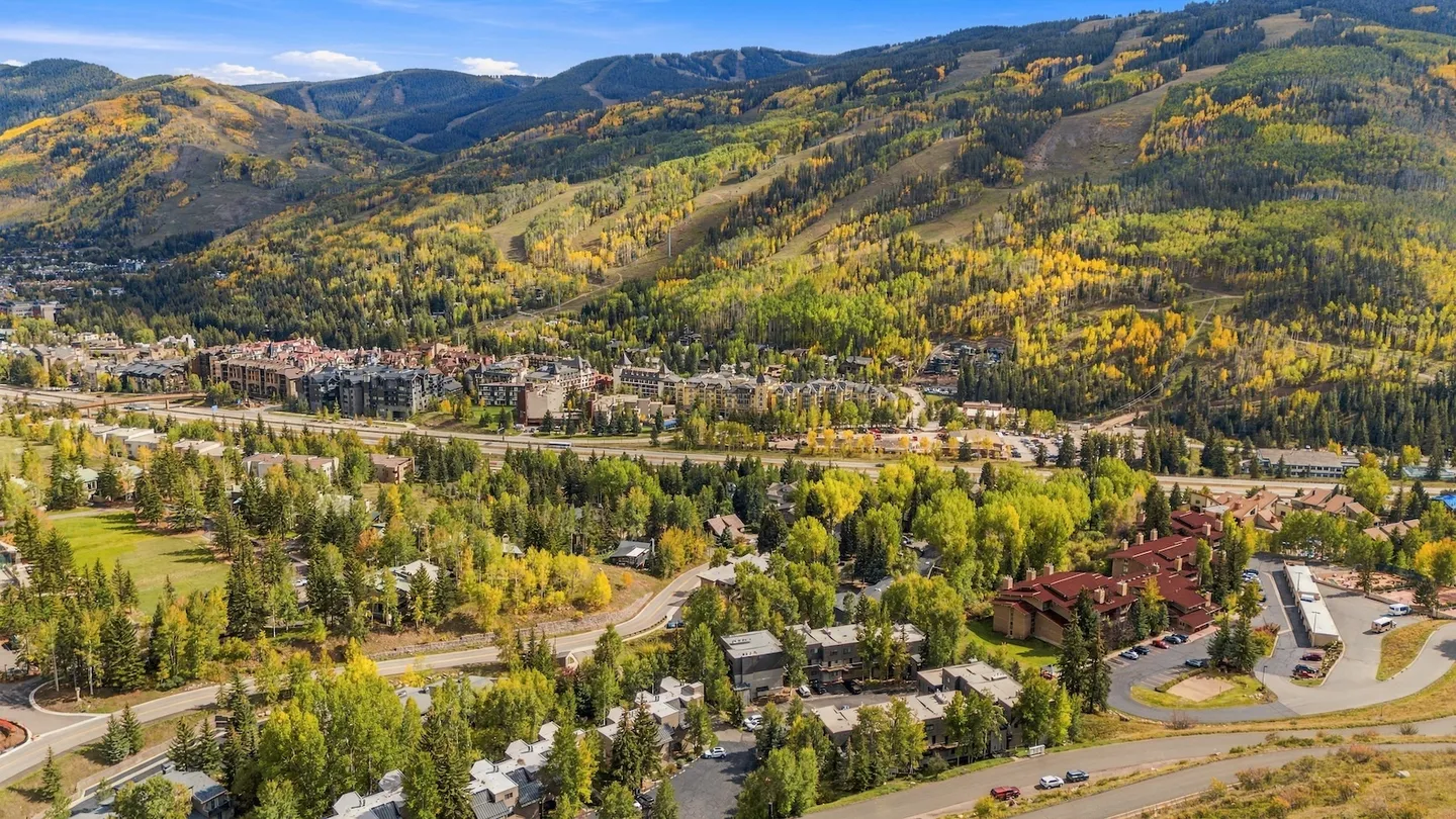 Aerial view of a mountain town and resort area surrounded by autumn foliage and tree-covered hills under a blue sky.