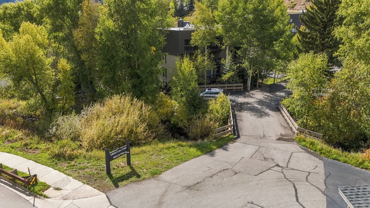 Elevated view of a paved driveway leading over a small bridge toward a multi-story building surrounded by dense trees.