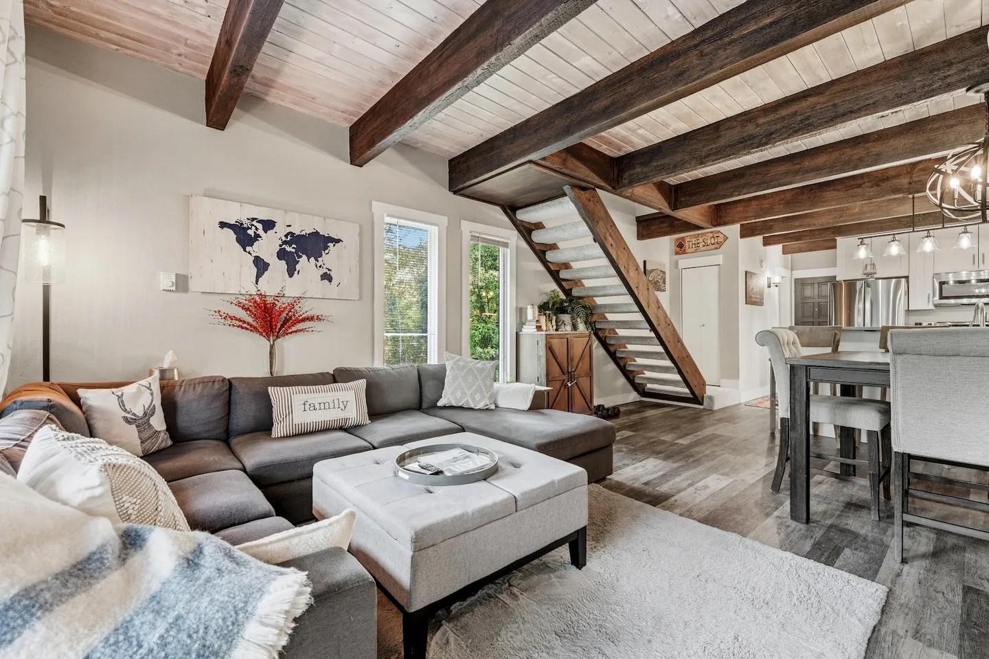 Open-concept living room with an L-shaped sectional sofa, tufted ottoman, exposed ceiling beams, and open-tread stairs leading upstairs.