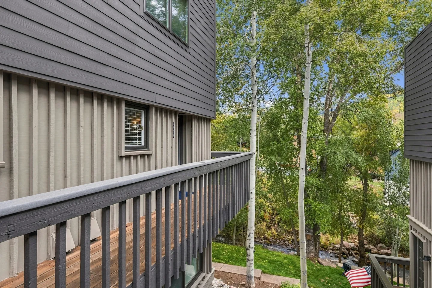 Exterior wooden balcony overlooking tall birch trees, greenery, and a nearby stream.