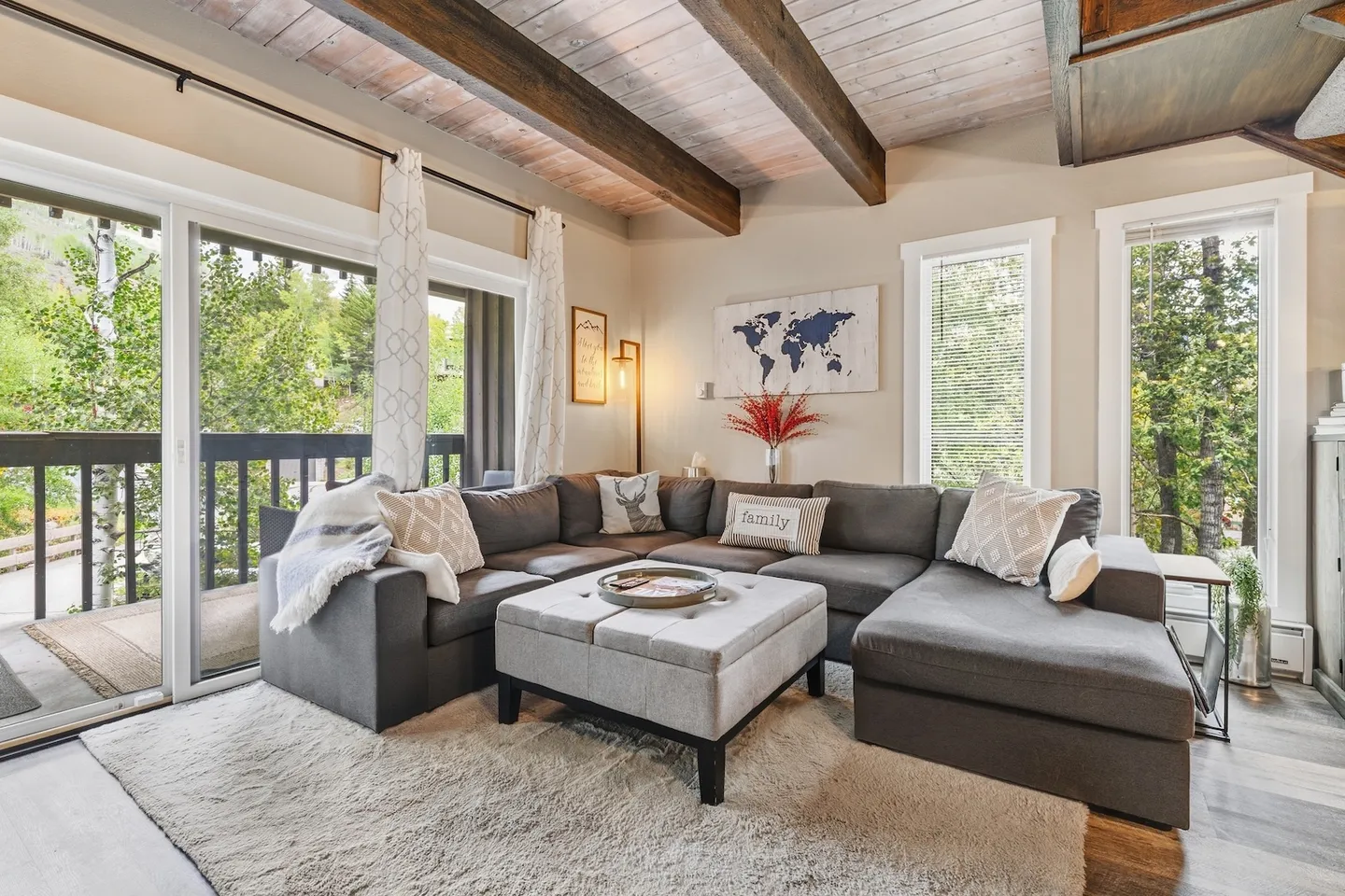 Living room with a large gray sectional sofa, an ottoman, wood-beamed ceiling, and sliding glass doors leading to a balcony.