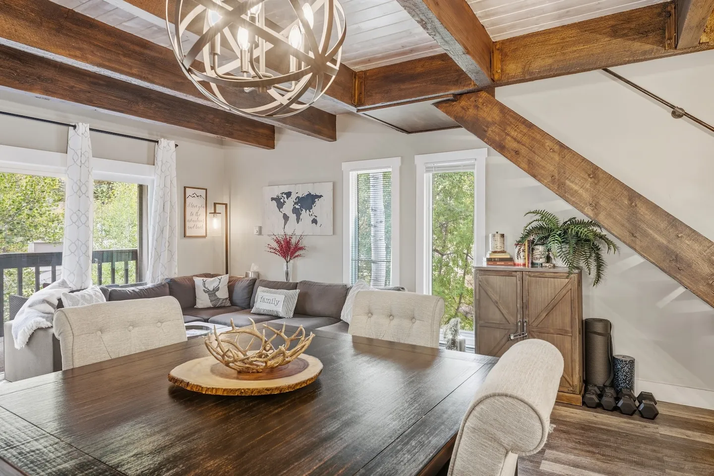 Dining area with a wooden table and upholstered chairs looking into an open living room with exposed ceiling beams and a staircase.