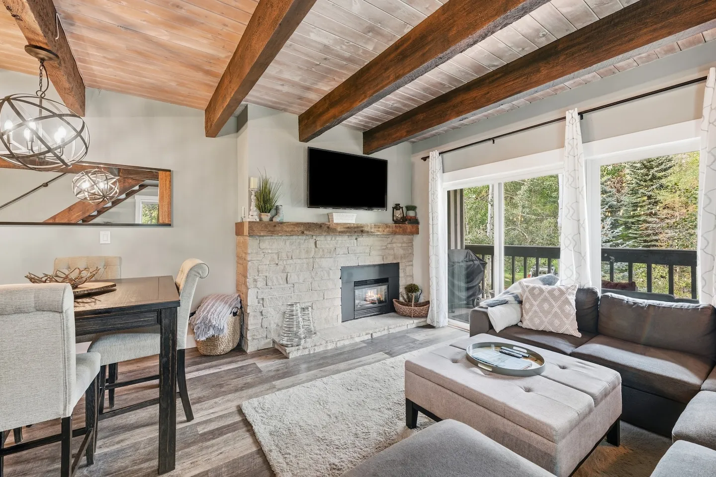 Living and dining area featuring exposed wood ceiling beams, a stone fireplace with a mounted TV, and sliding glass doors leading to a balcony.