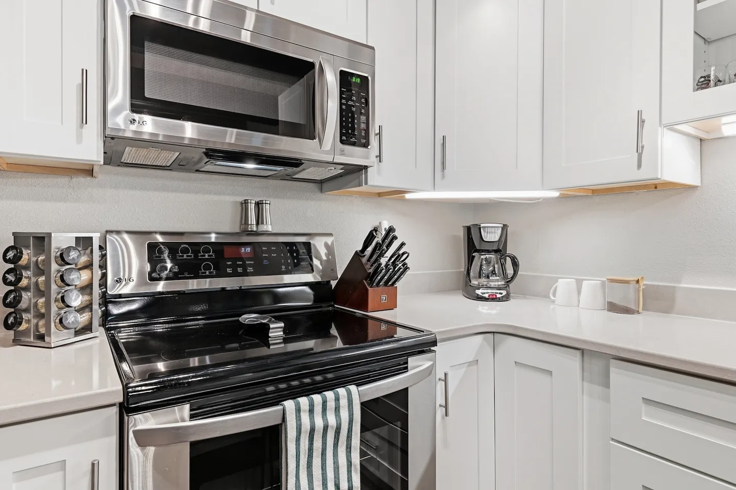 Kitchen counter area with stainless steel stove, over-the-range microwave, spice rack, knife block, and coffee maker against white cabinetry.