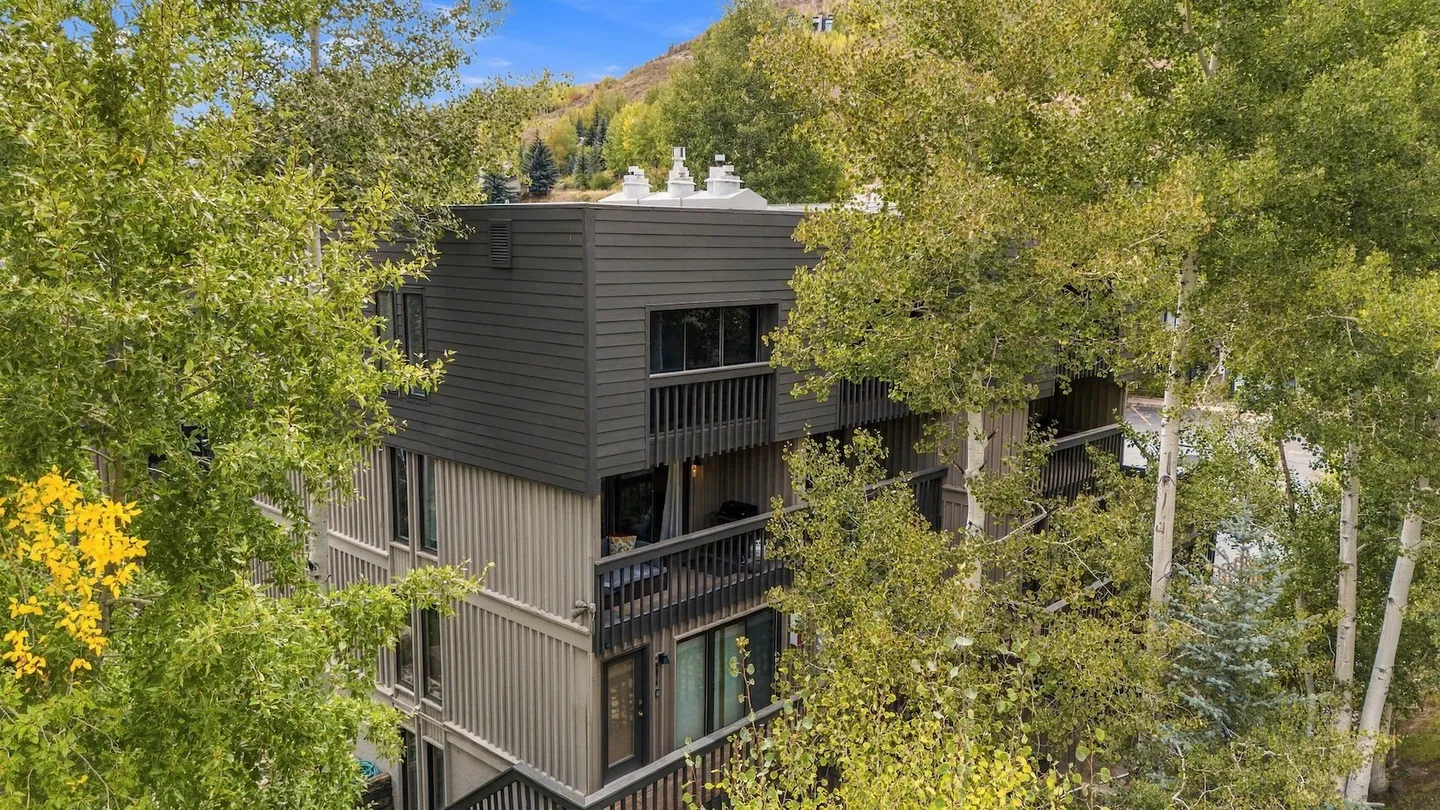 Exterior view of a multi-story building with dark and tan siding, balconies, and surrounding green and yellow trees under a blue sky.
