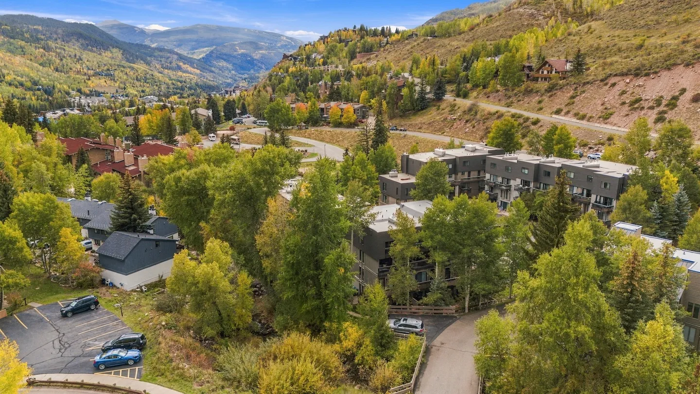 Aerial view of a mountain valley community with multi-story buildings, paved roads, and green and yellow trees.