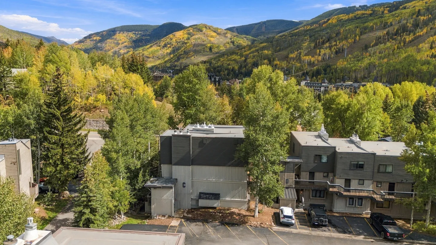 Elevated view of multi-story townhomes nestled among green and yellow trees with rolling mountains in the background under a blue sky.