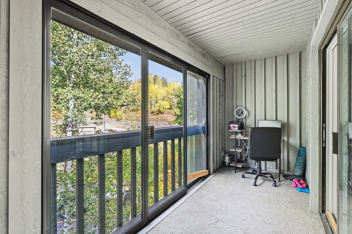 Covered balcony or sunroom with large glass sliding doors and an office desk setup overlooking outdoor foliage.