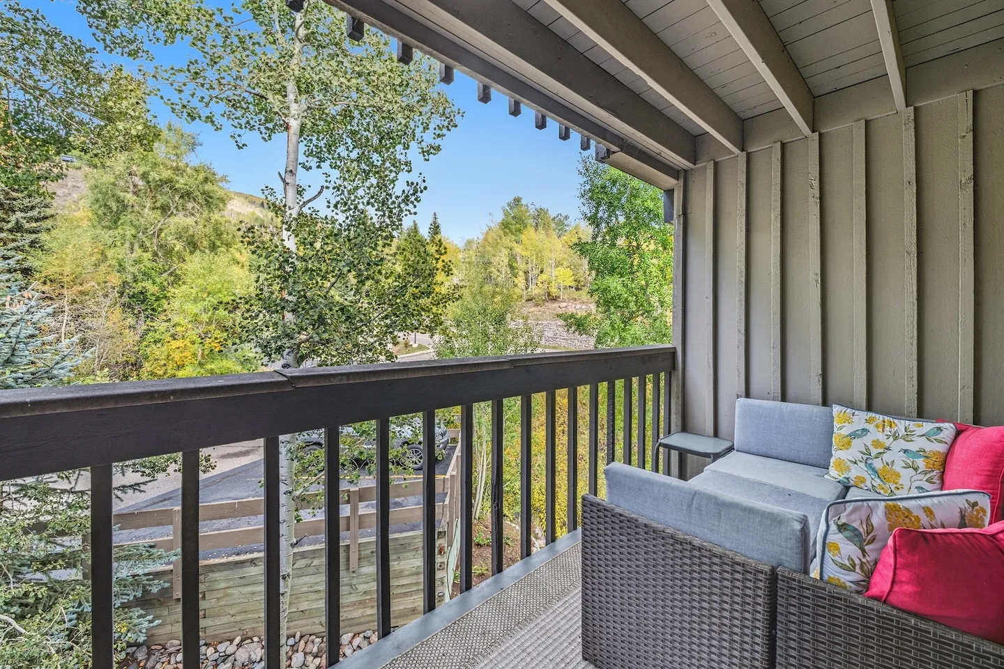 A covered balcony with an outdoor wicker sofa and cushions overlooking trees under a clear blue sky.