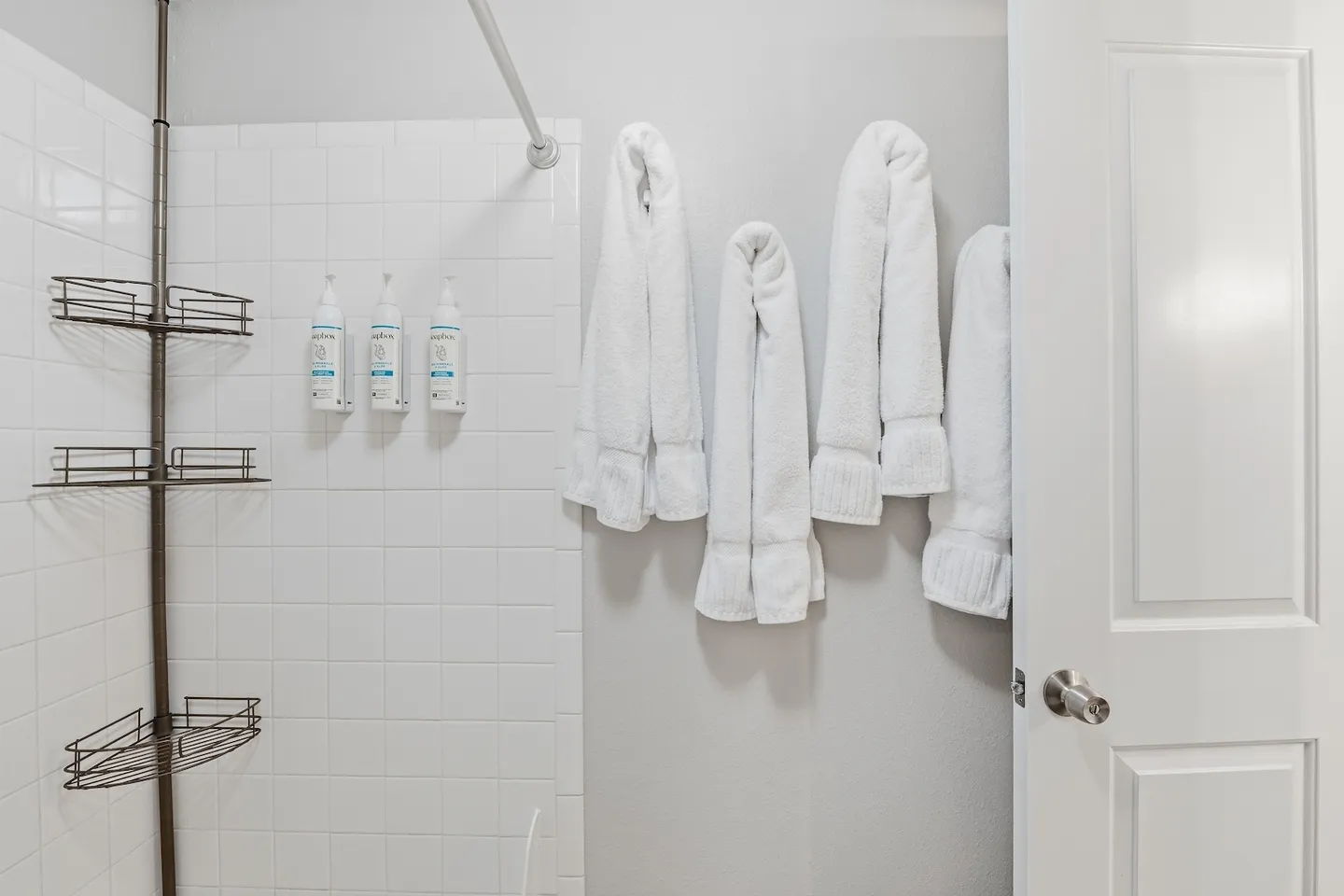 Bathroom showing white subway-tiled shower area with wire shelving and wall-mounted toiletries next to hanging white towels.