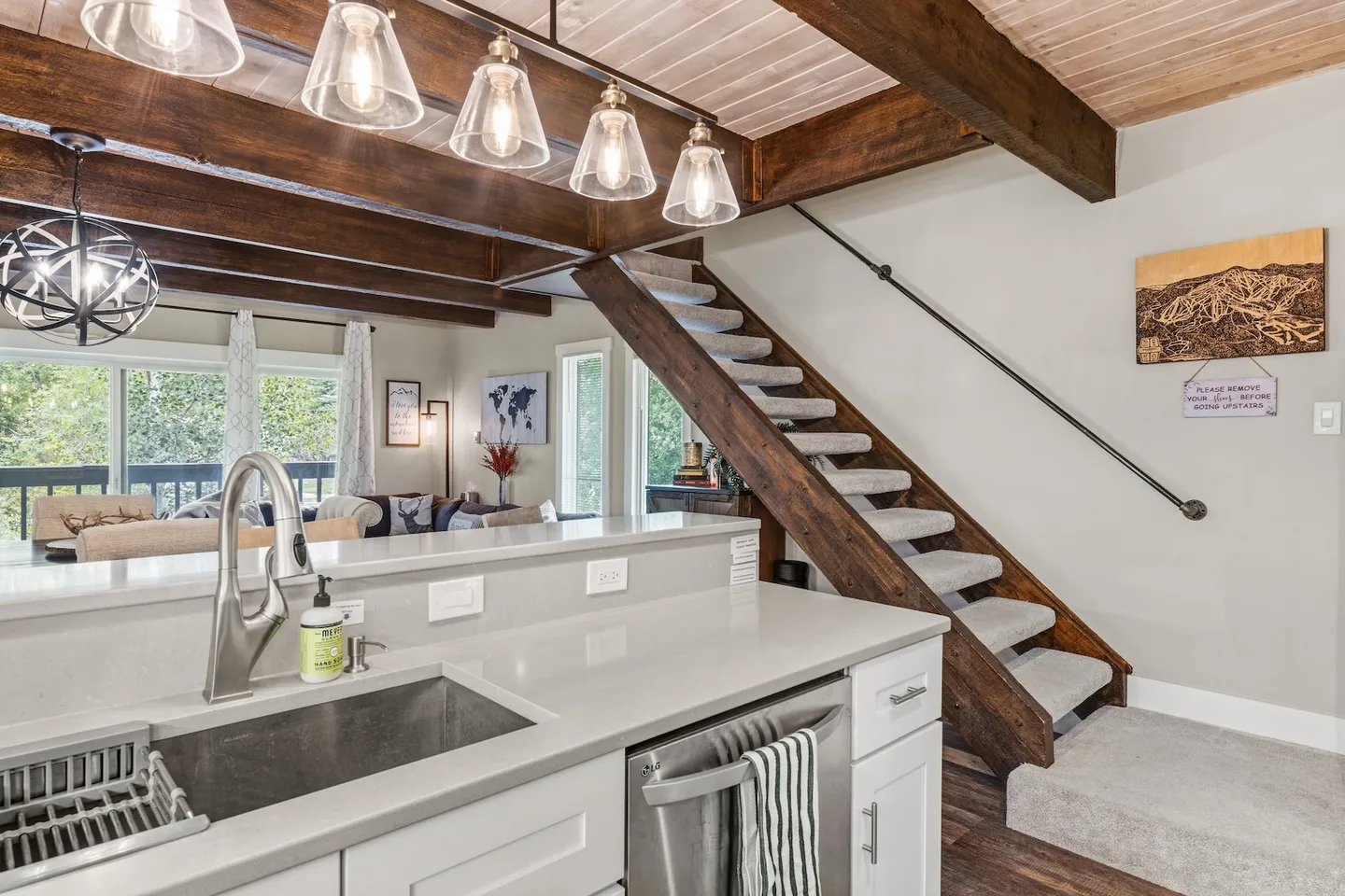A kitchen counter with an undermount sink and stainless steel dishwasher next to a wooden staircase leading upstairs in an open-concept living area.