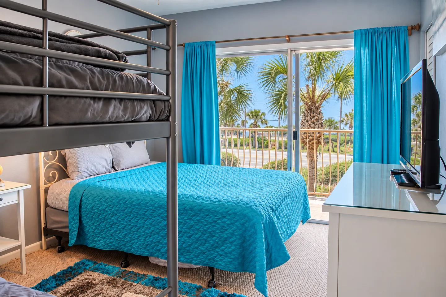 Bedroom with a bunk bed, a double bed with a bright blue coverlet, a TV console, and sliding glass doors to a balcony with palm trees.