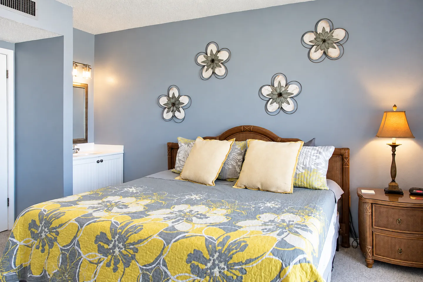 Bedroom featuring a made bed with yellow and grey floral bedding, blue walls with flower wall art, and an adjacent vanity sink.