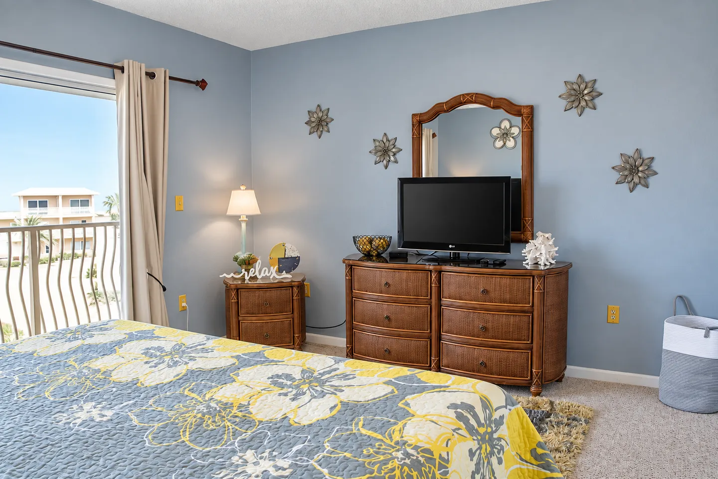 Bedroom with blue walls, a bed with a floral quilt, a wicker dresser with a television, and a sliding glass door leading to a balcony.