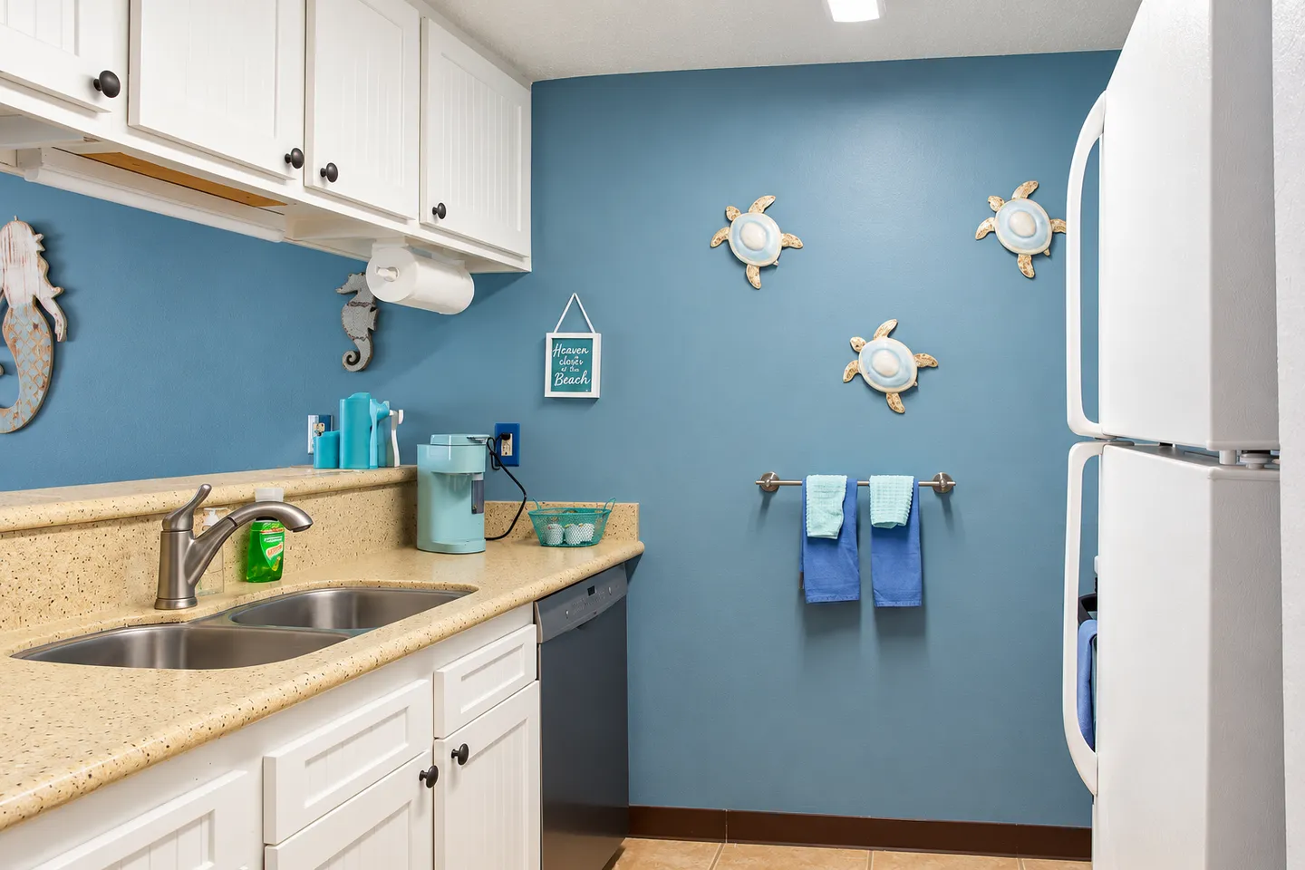 Kitchen area featuring blue accent walls decorated with sea turtle and seahorse wall art, white cabinets, a double sink, and a white refrigerator.