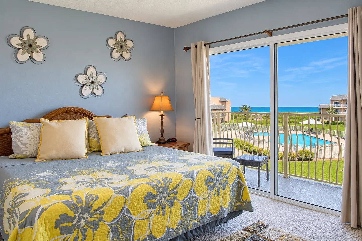 Bedroom with blue walls, floral bedspread, and sliding glass doors leading to a balcony with views of a pool and the ocean.