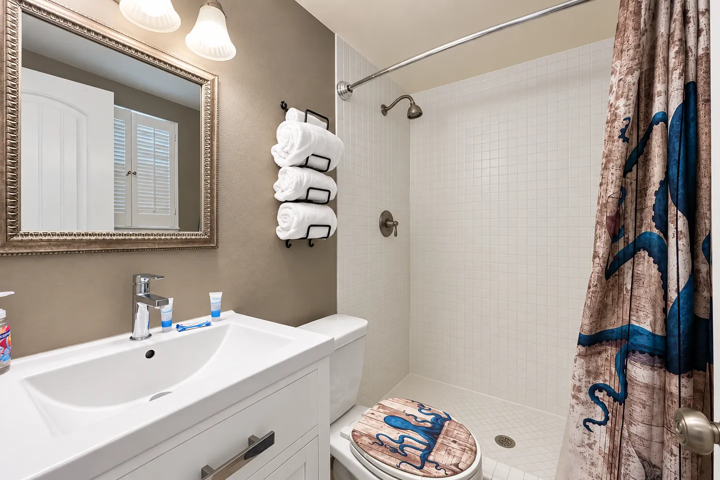 Bathroom with white vanity, framed mirror, toilet with decorated lid, and tiled walk-in shower with curtain.