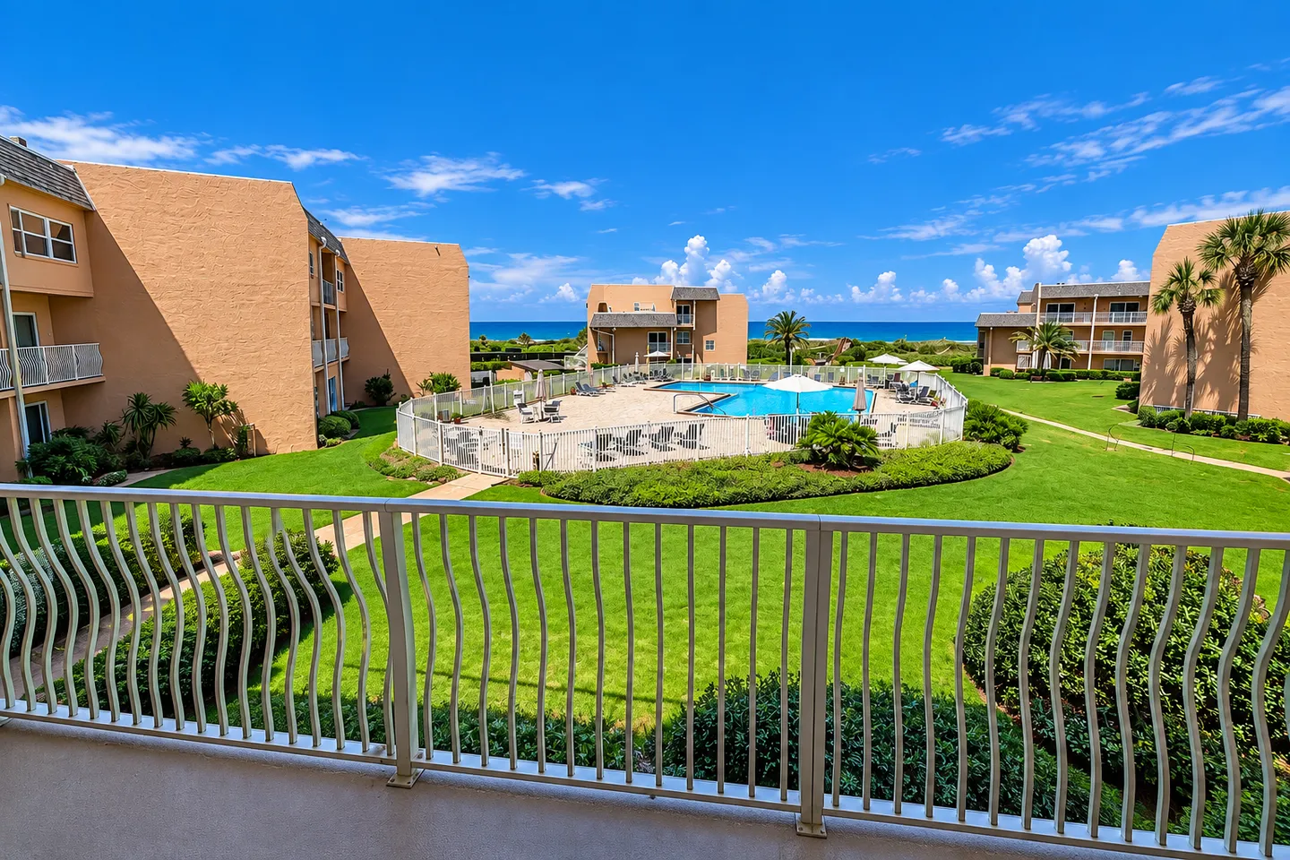 Balcony view overlooking a fenced community swimming pool, manicured lawns, multi-story buildings, and the ocean under a blue sky.