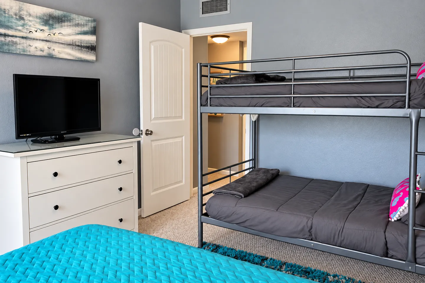 Bedroom featuring a metal bunk bed dressed in grey, a white dresser with a television, and a doorway leading into a hallway.