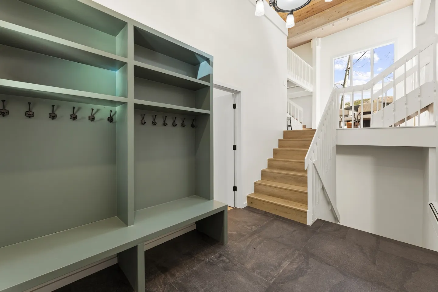 Entryway mudroom area featuring built-in sage green cubbies with coat hooks, bench seating, dark floor tiles, and stairs leading up to an open upper level.