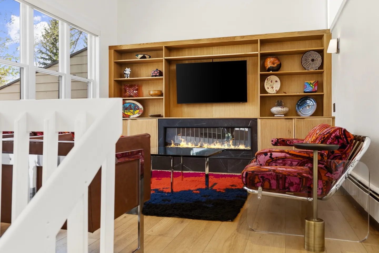Living area featuring a modern linear fireplace beneath a mounted television within built-in wooden shelving, accented by a colorful patterned armchair and rug.