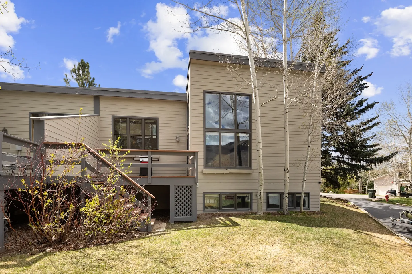 Exterior rear view of a two-story tan-sided home with a raised wooden deck and large windows under a blue sky.
