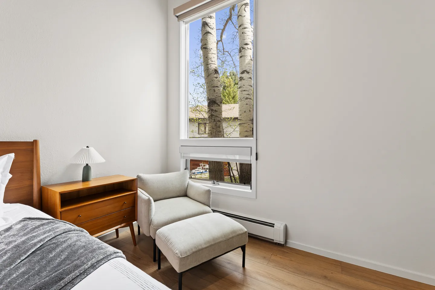Bedroom corner with an armchair and ottoman placed next to a tall window showing birch trees outside.