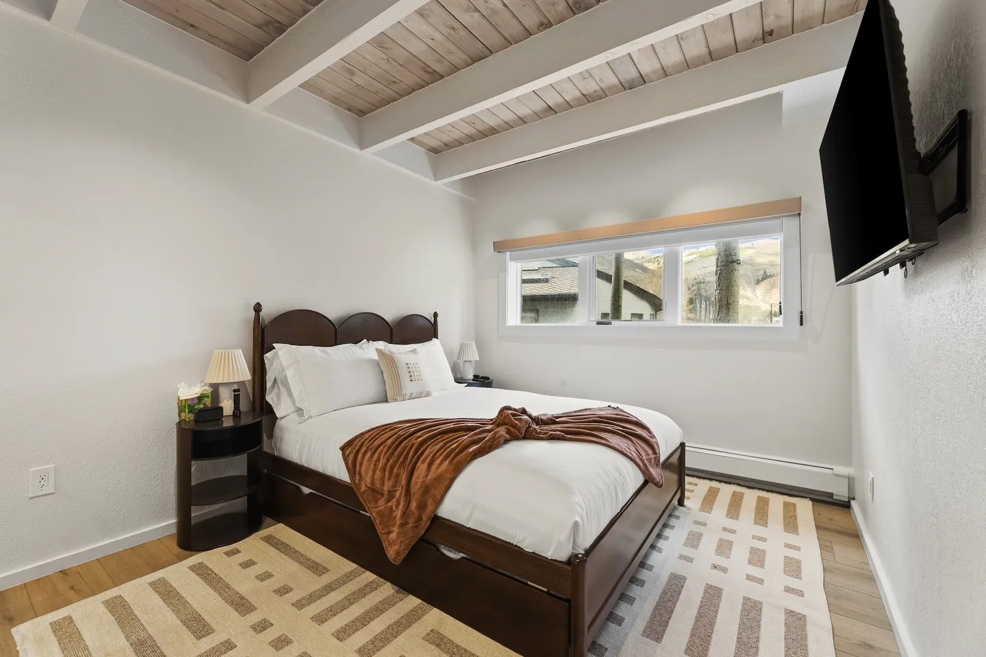 Bedroom with a wood-frame bed, exposed beam ceiling, narrow horizontal window, and wall-mounted television.