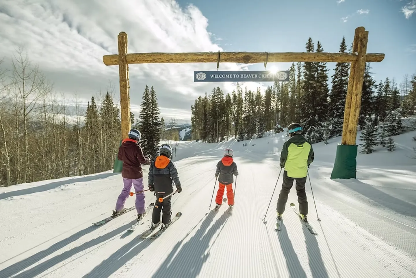Four skiers viewed from behind on a groomed snow run approaching a wooden archway with a Welcome to Beaver Creek sign.