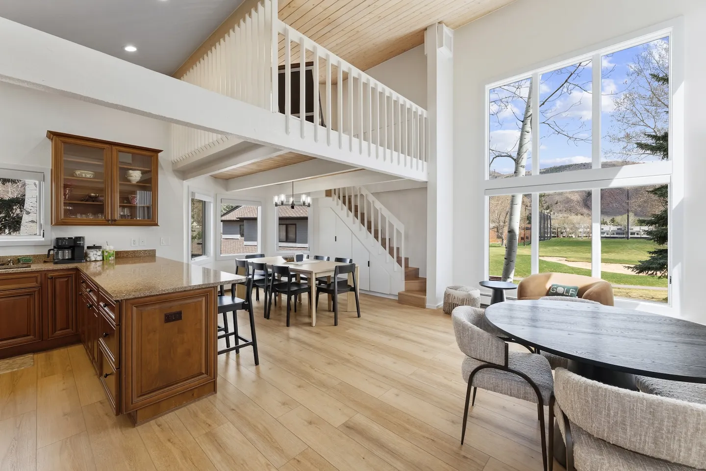 Open-concept interior with a kitchen island, dining areas, vaulted wood ceiling, loft, and large two-story window overlooking a grassy landscape.