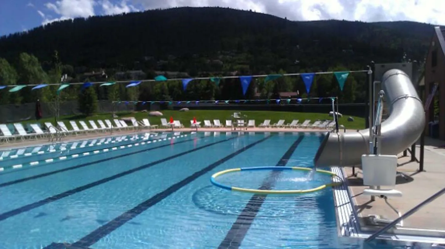 Large outdoor swimming pool with lap lanes, a water slide, and rows of lounge chairs against a backdrop of green mountains.