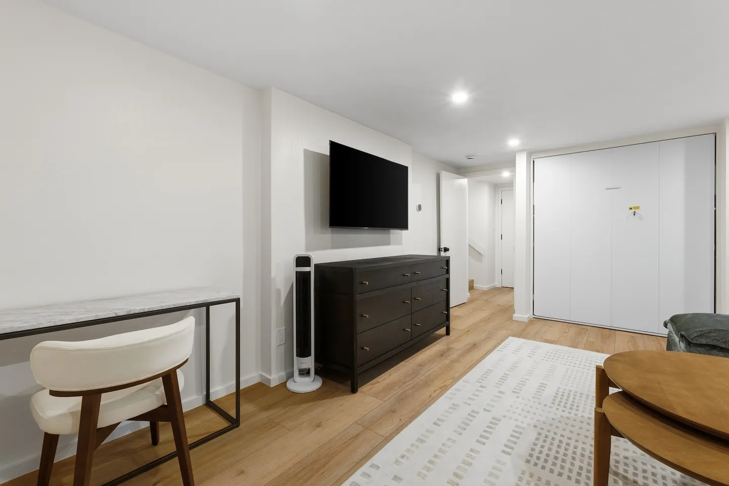 A modern interior room with wood-style flooring, a desk and chair, a dark dresser beneath a wall-mounted TV, and a closed Murphy bed unit.