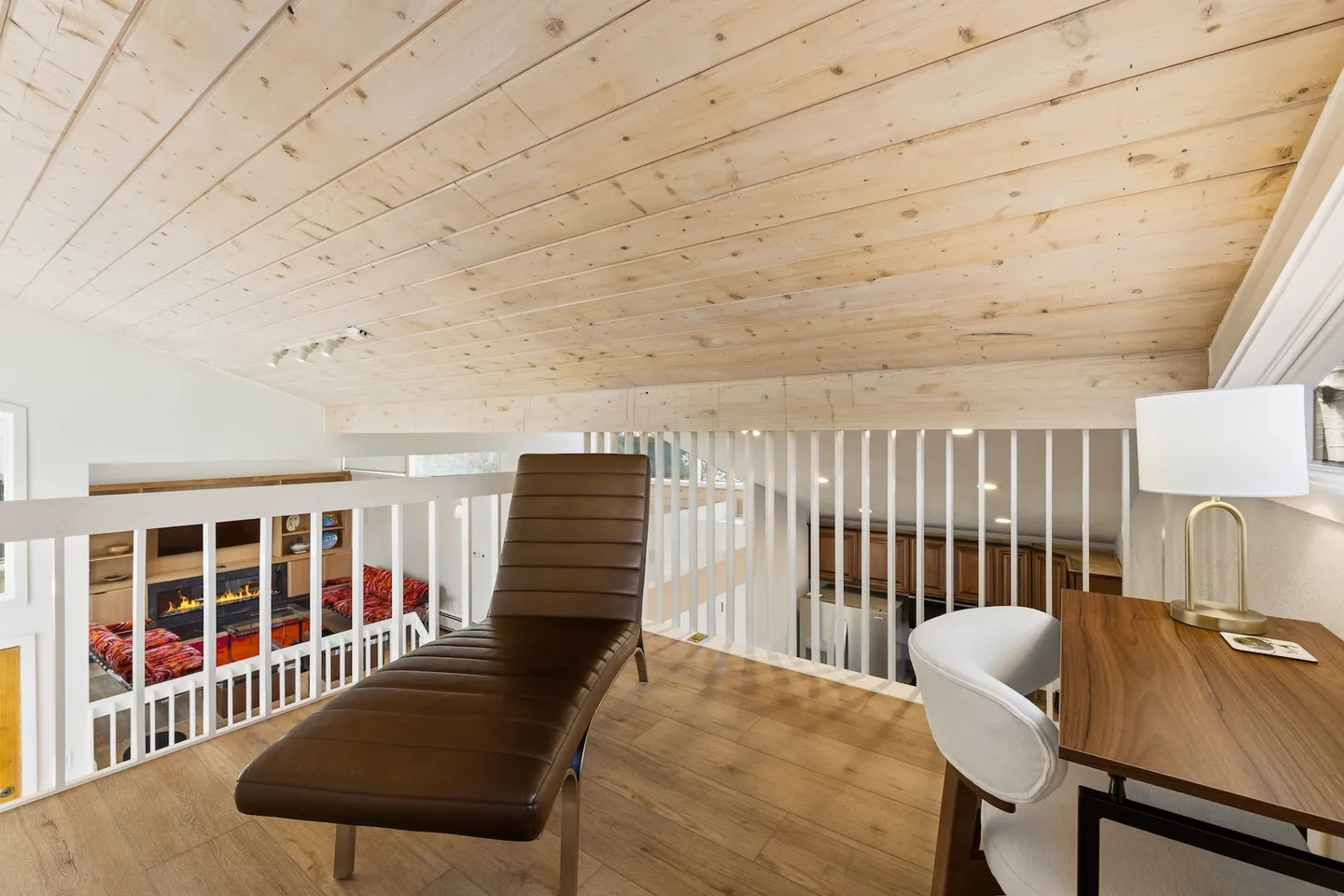 A modern loft space with sloped wood plank ceiling, brown leather chaise lounge, desk, and view over white railing to the lower level.