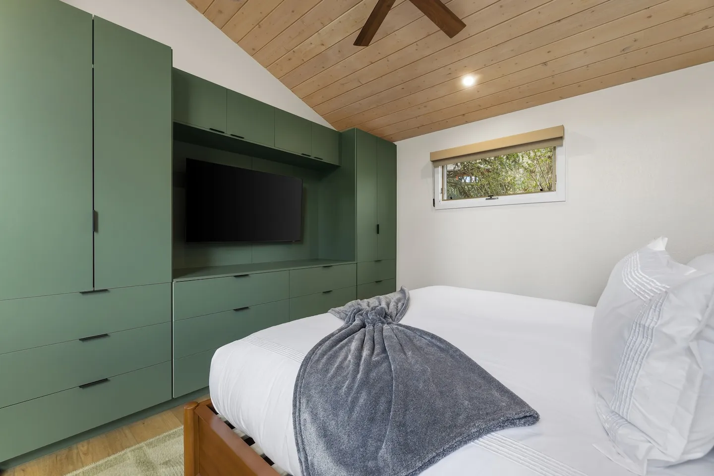 Bedroom with a vaulted wood-paneled ceiling, a bed made with white linens, and custom green built-in cabinetry surrounding a wall-mounted TV.
