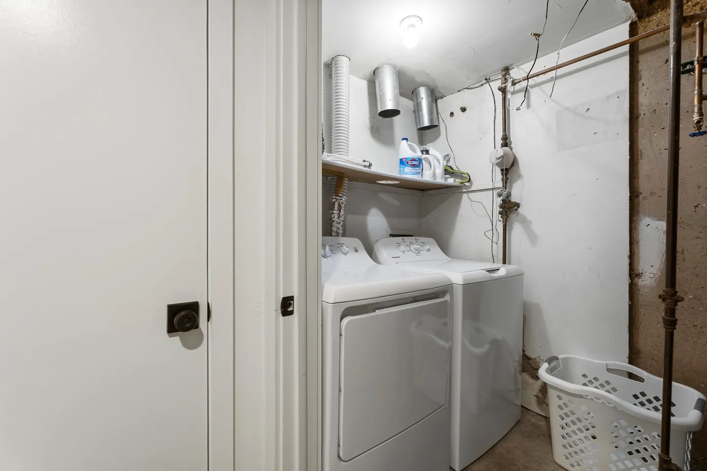 A utility closet or basement laundry area with a white washer and dryer, laundry basket, overhead shelf, and exposed pipes.