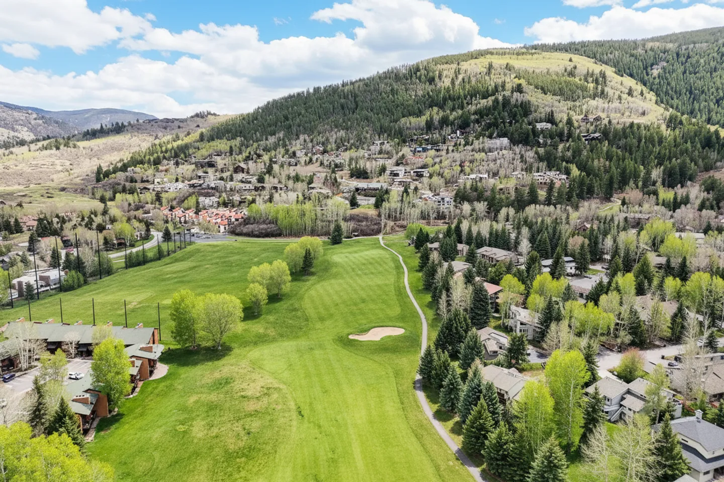 Aerial view of a golf course fairway bordered by trees, paved paths, and mountain residences under a partly cloudy sky.
