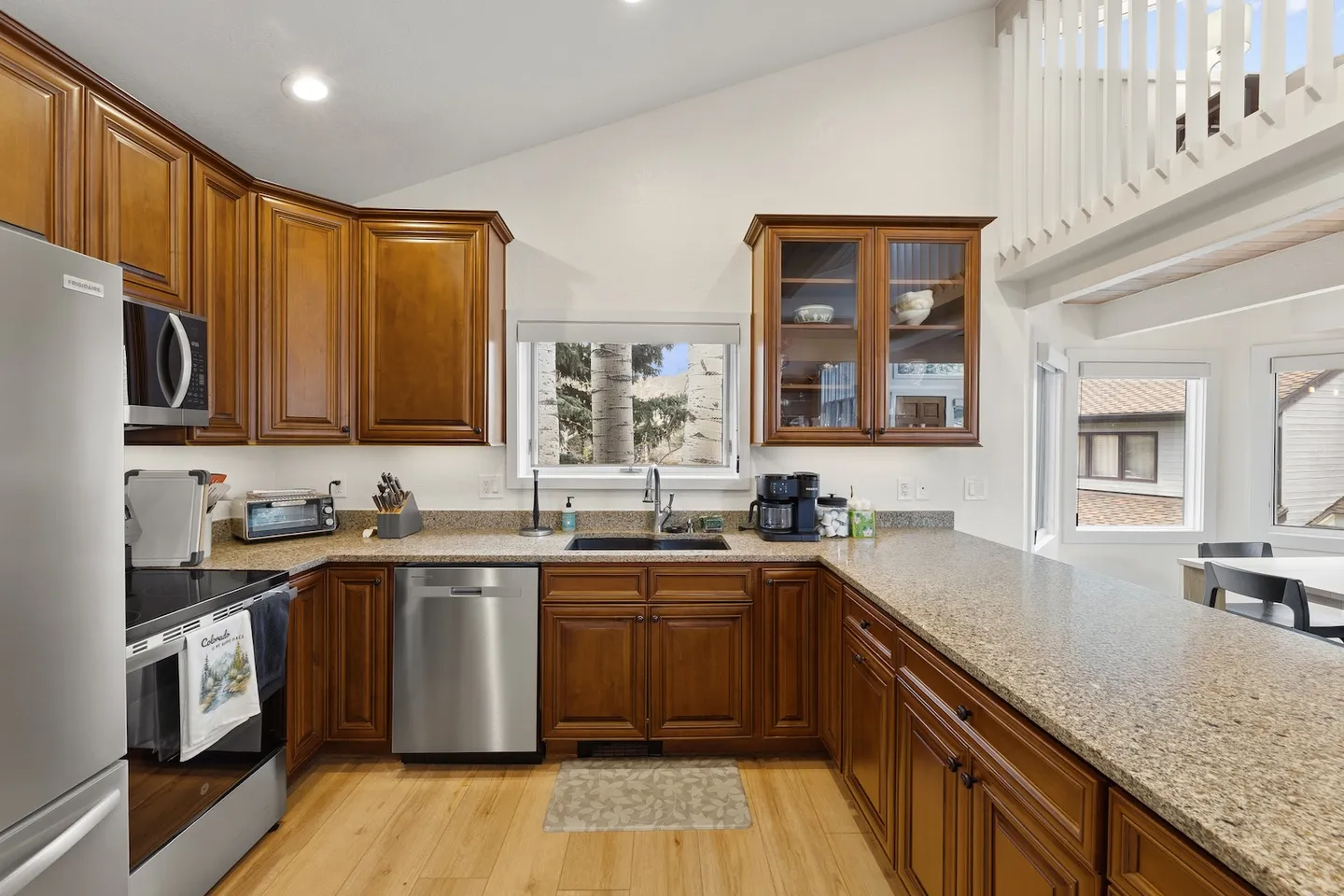 Kitchen with wooden cabinetry, speckled countertops, stainless steel appliances, and a window above the sink looking out at trees.