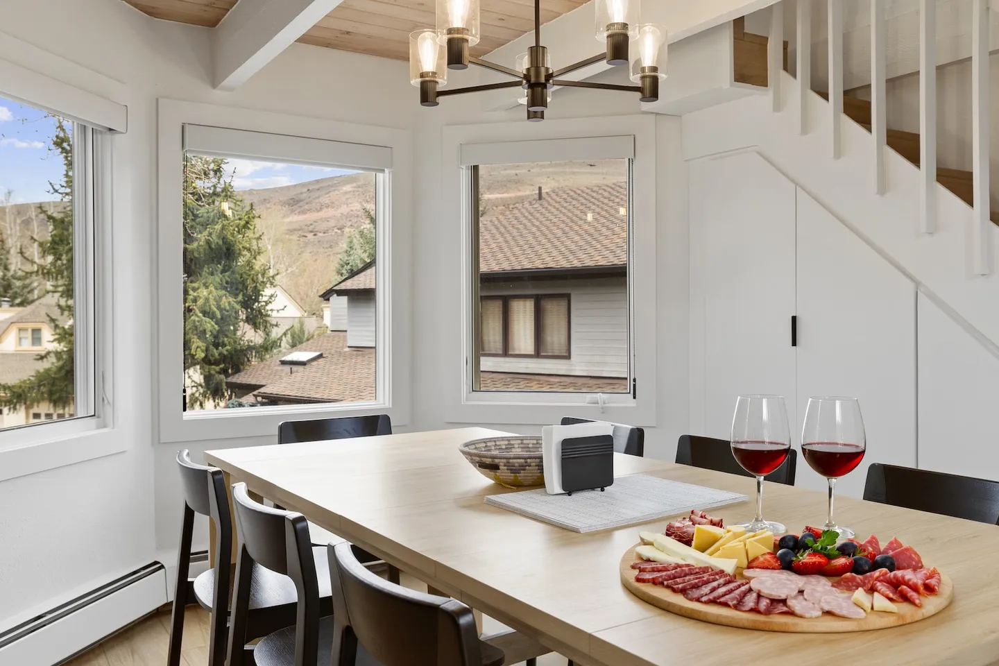 Dining table set with a charcuterie board and wine glasses near corner windows overlooking hills.