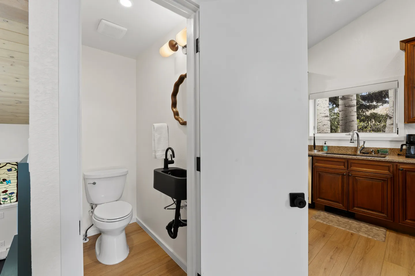 Powder room viewed through an open white door, featuring a white toilet, wall-mounted dark sink, and mirror, adjacent to a kitchen with wood cabinets.