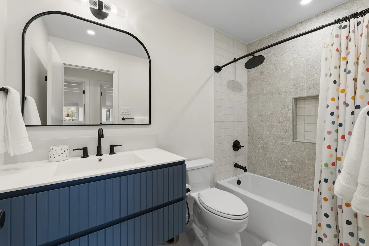 Bathroom with a blue slatted vanity, arched mirror, black fixtures, toilet, and combination bathtub and shower with polka dot curtain.