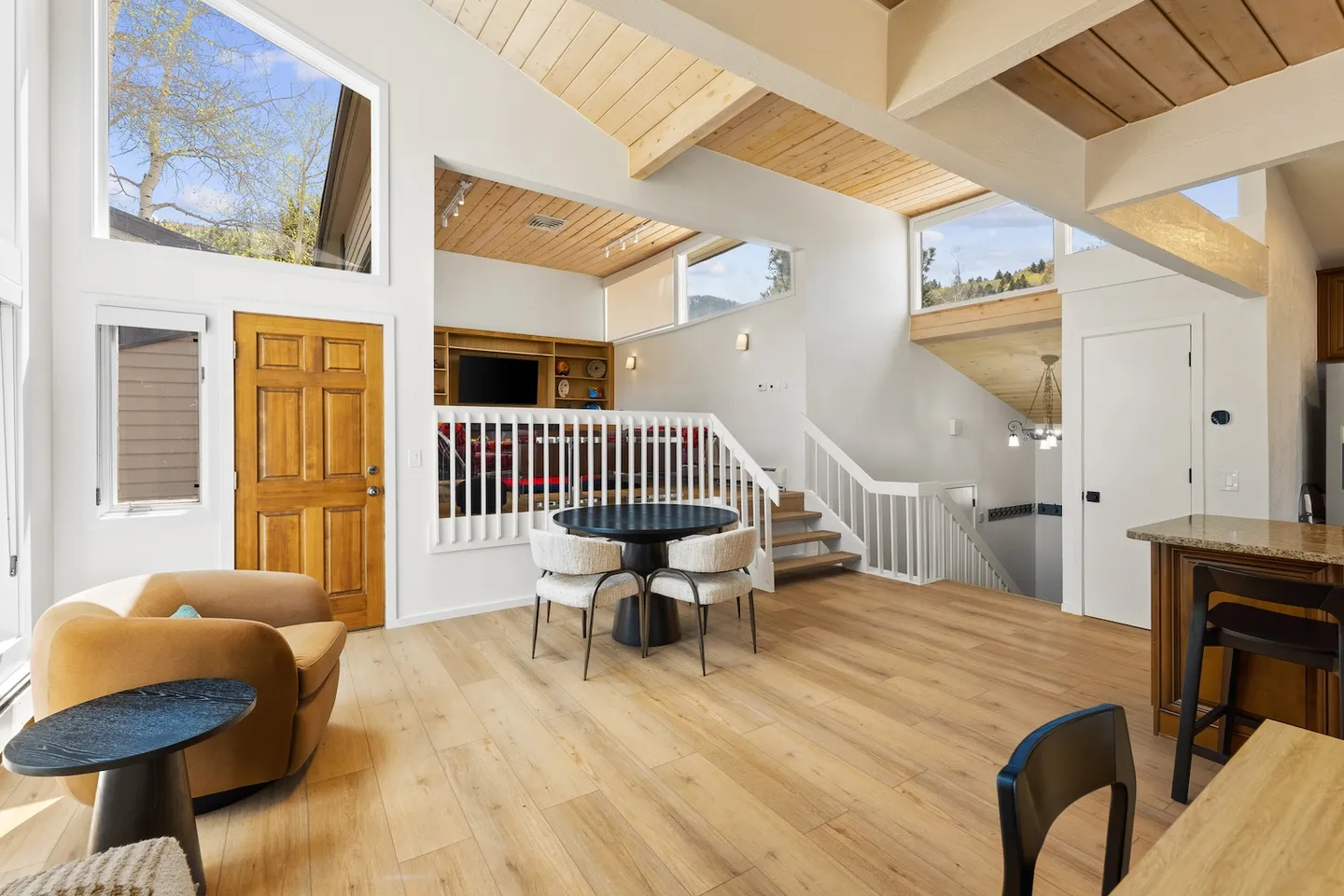 Open-concept living space with high ceilings, exposed wood beams, light wood floors, an entryway door, and stairs leading to split levels.