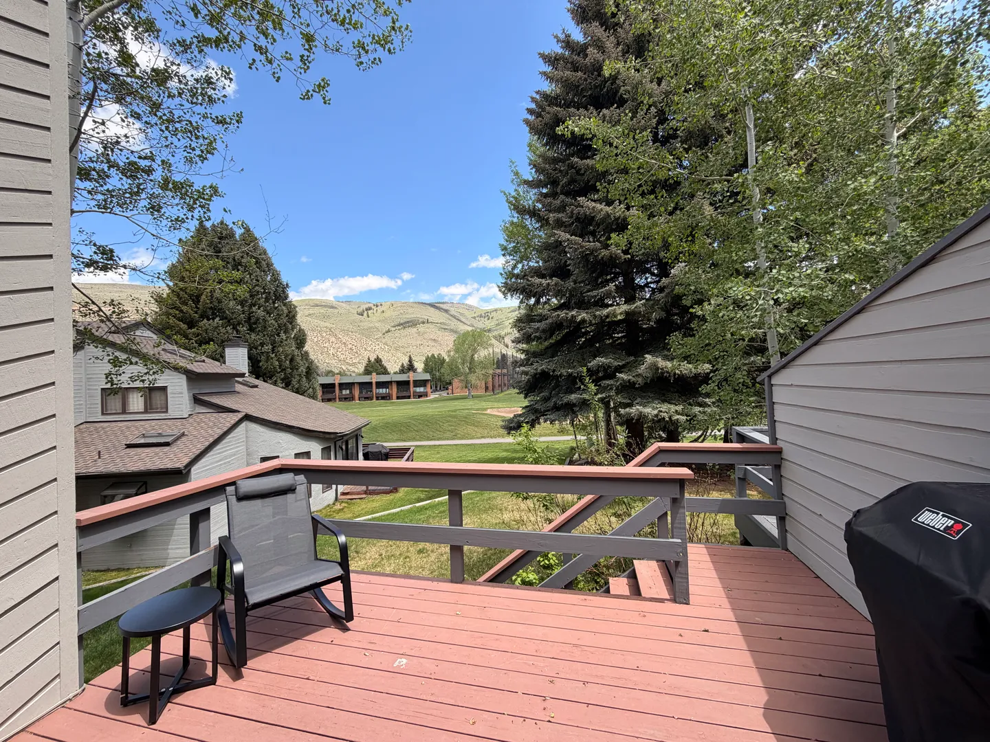 Elevated wooden deck with a rocking chair, small side table, and covered grill overlooking green hills and nearby houses.