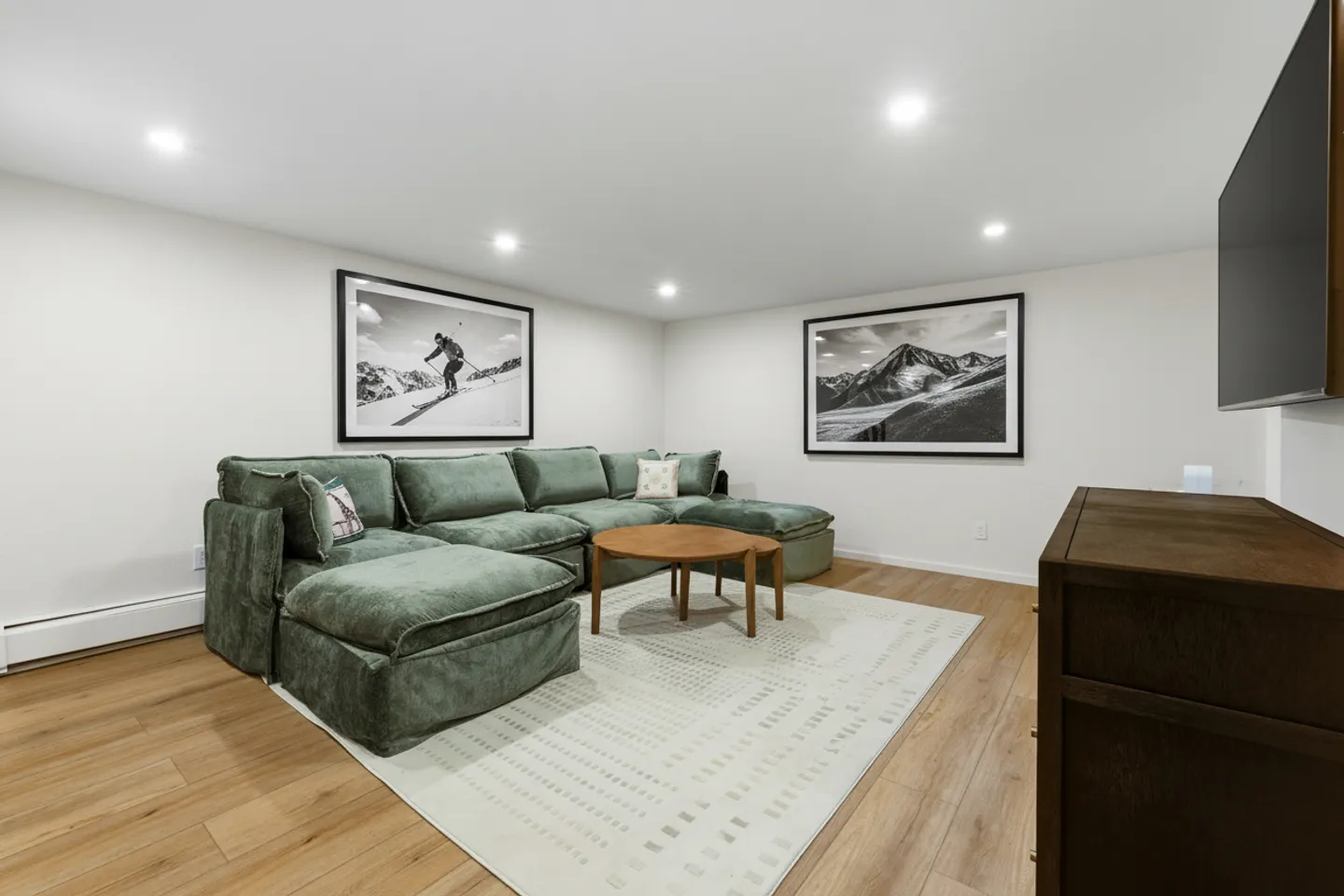 A living room with a large green sectional sofa, wooden coffee table, area rug, and framed mountain art prints.