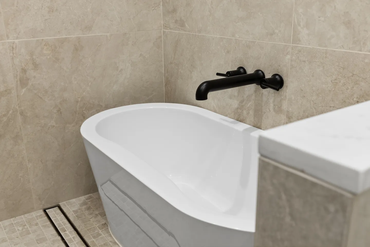 A close-up view of a modern white soaking tub with a wall-mounted matte black faucet and neutral tile walls.