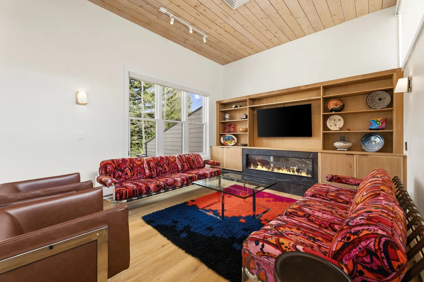 Living room with patterned red sofas, wood-paneled ceiling, large area rug, and built-in shelving with an electric fireplace and TV.