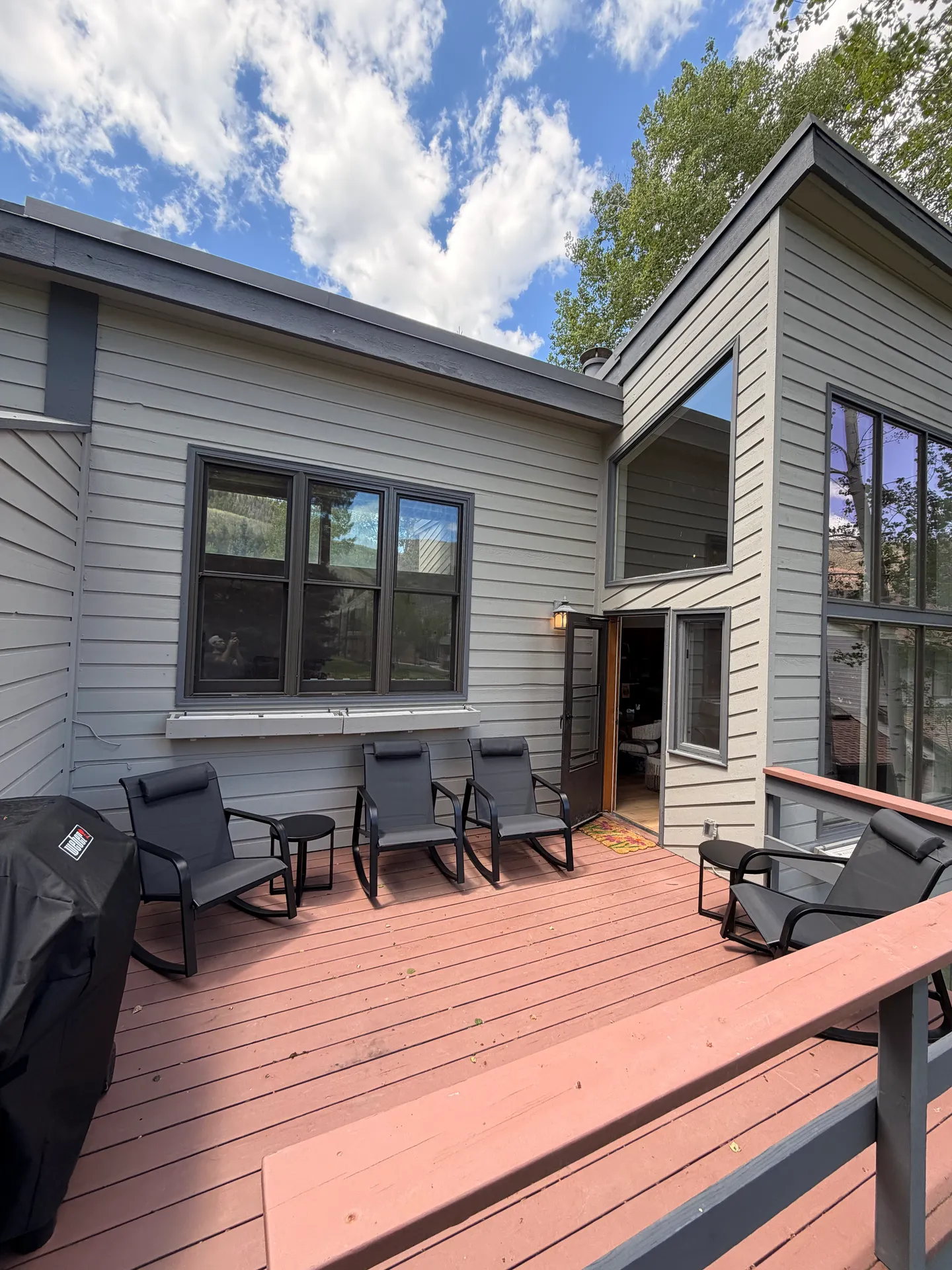 Outdoor wood deck with several chairs, small side tables, and a covered grill next to a gray-sided house under a partly cloudy sky.