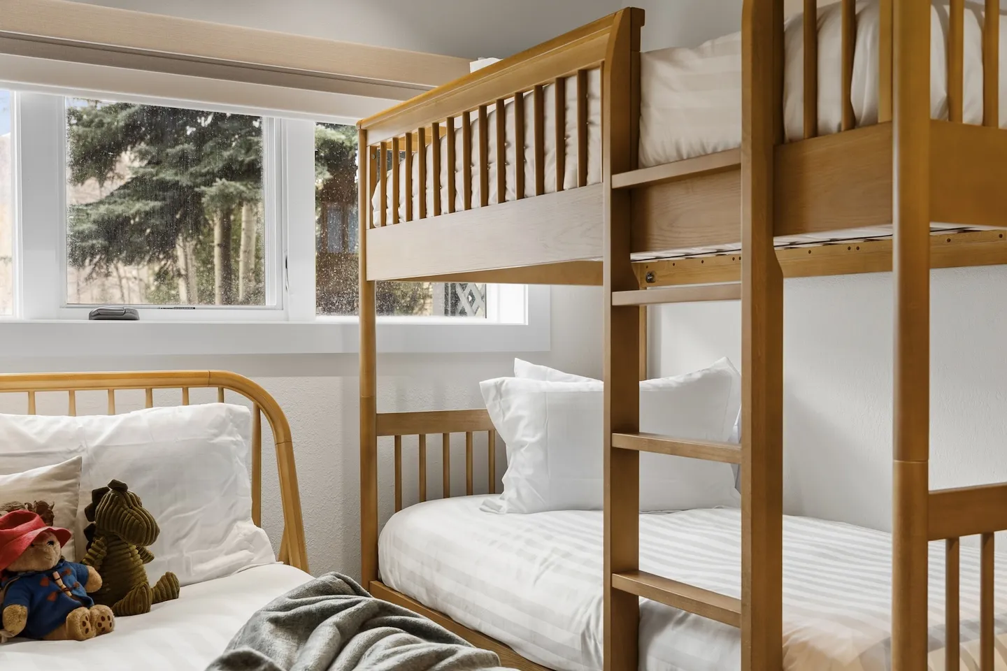 A bedroom corner featuring a wooden bunk bed and a separate single bed with stuffed animals next to a bright window.