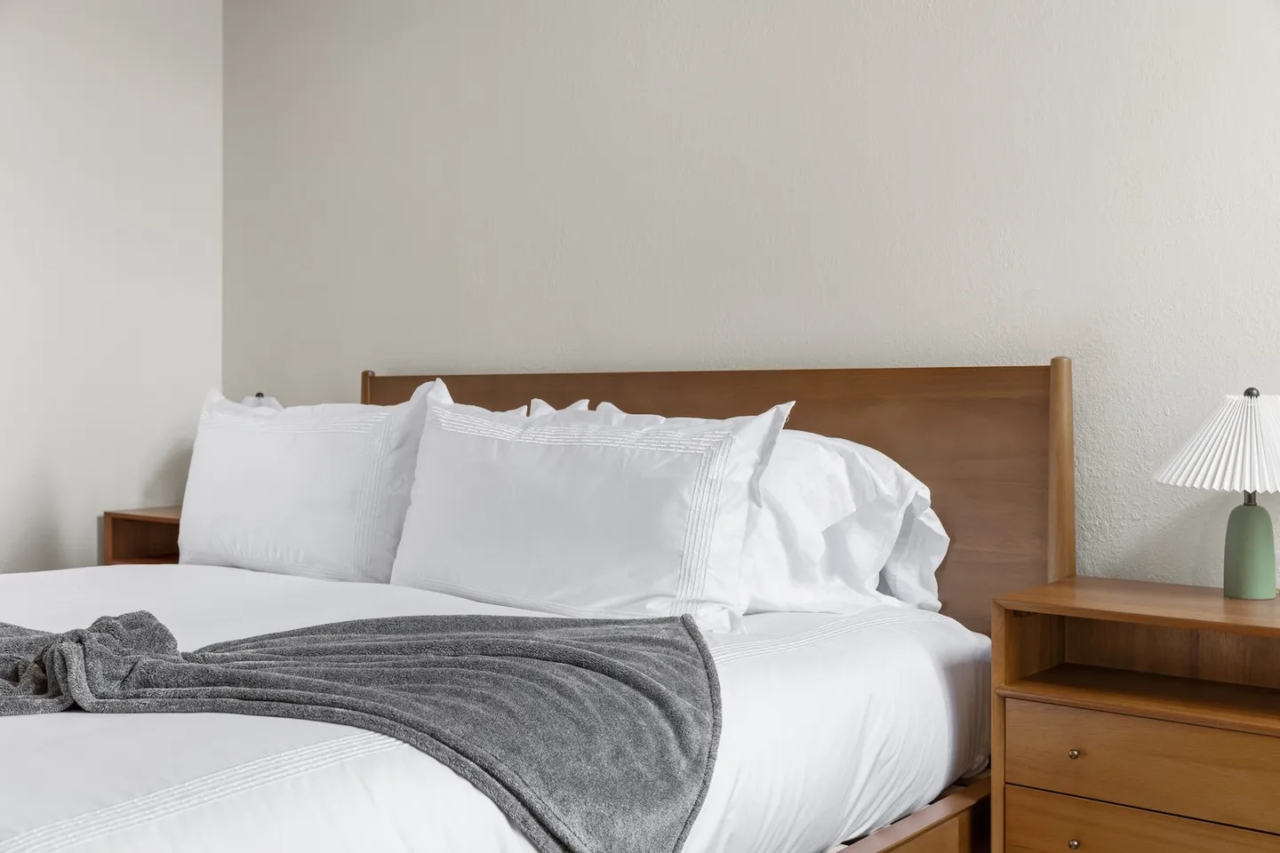 A made bed with white linens and a gray throw blanket flanked by wooden nightstands against plain walls.